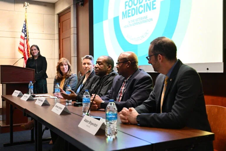 a panel discussion on “Food is Medicine in the Veterans Health Administration” in Washington, DC. Six speakers sit at a long table with microphones and water bottles, while a moderator stands at a podium beside an American flag. A large screen behind them displays the event title.