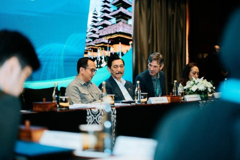 three panelists seated at a conference table during a presentation, with a screen displaying an Indonesian temple in the background