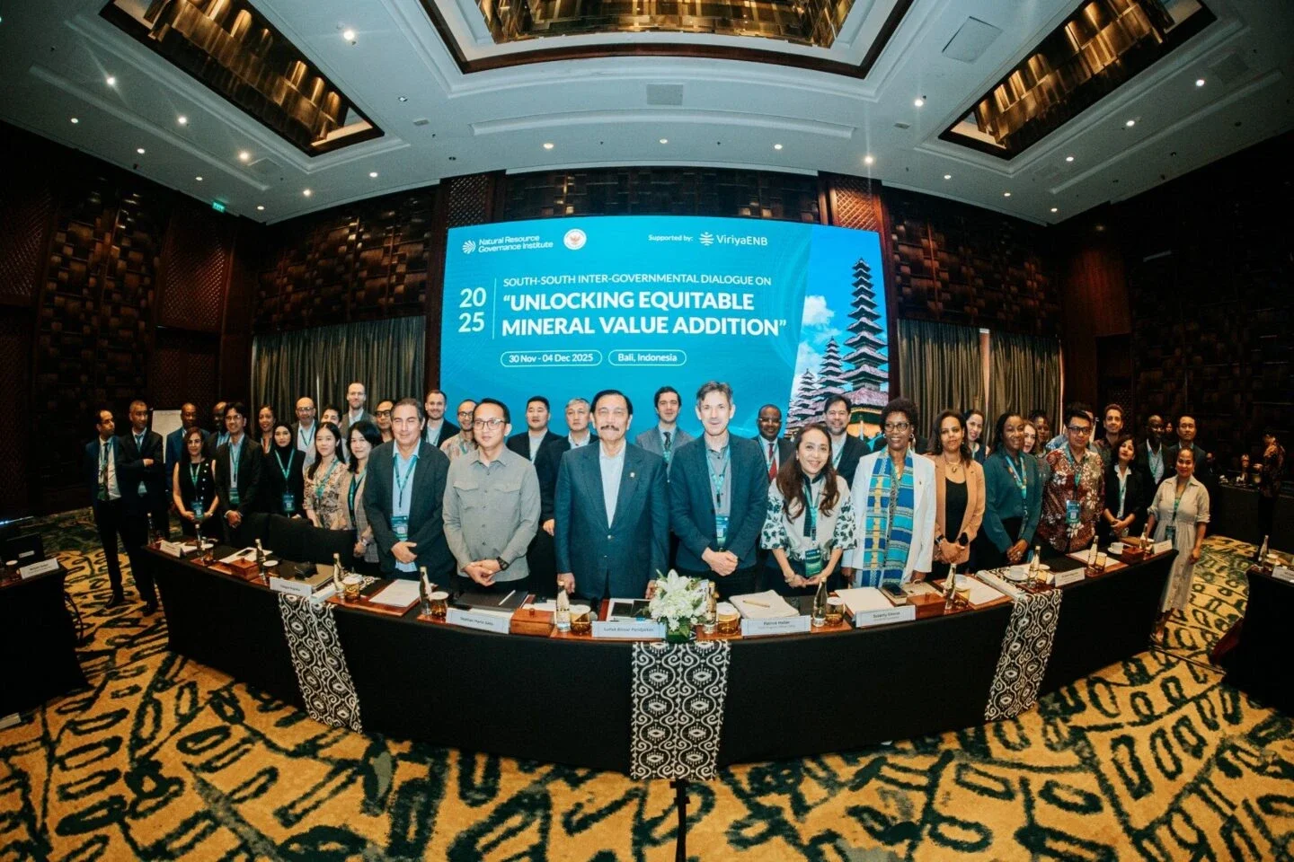group photo of conference participants standing in an elegant hotel ballroom beneath a large screen displaying 'Unlocking Equitable Mineral Value Addition' conference branding for an event in Bali, Indonesia