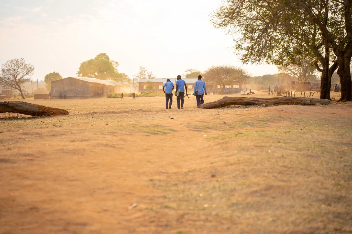 three people in blue work uniforms walking down a dusty dirt road in a rural African village during golden hour, with school buildings and community members visible in the hazy background