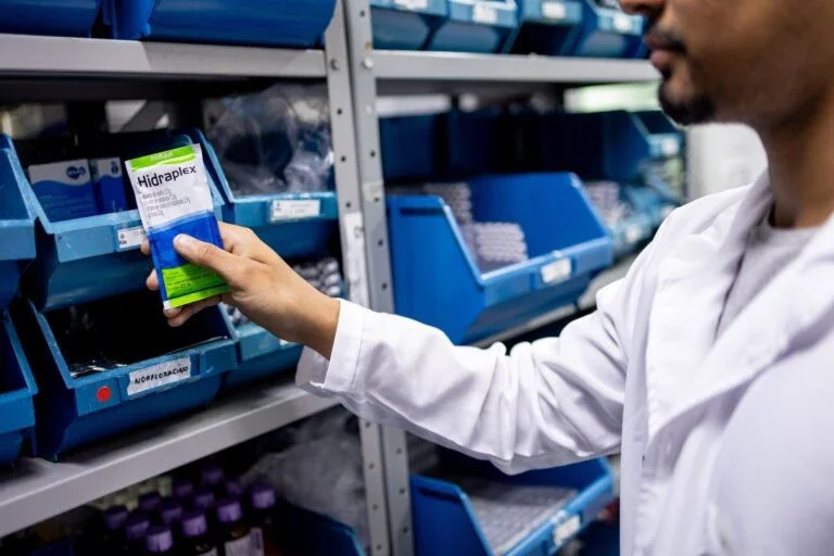 a healthcare worker in a white coat holds a box of Hidraplex medication while standing in front of organized blue storage bins on metal shelving