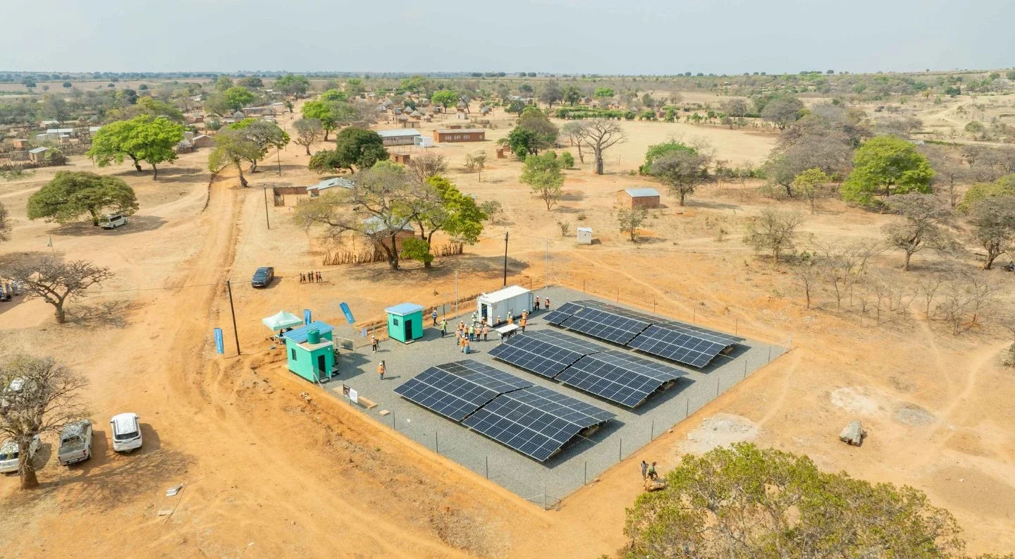 aerial view of a solar panel installation with turquoise support buildings in a rural African village, surrounded by scattered trees and dirt roads under a hazy sky